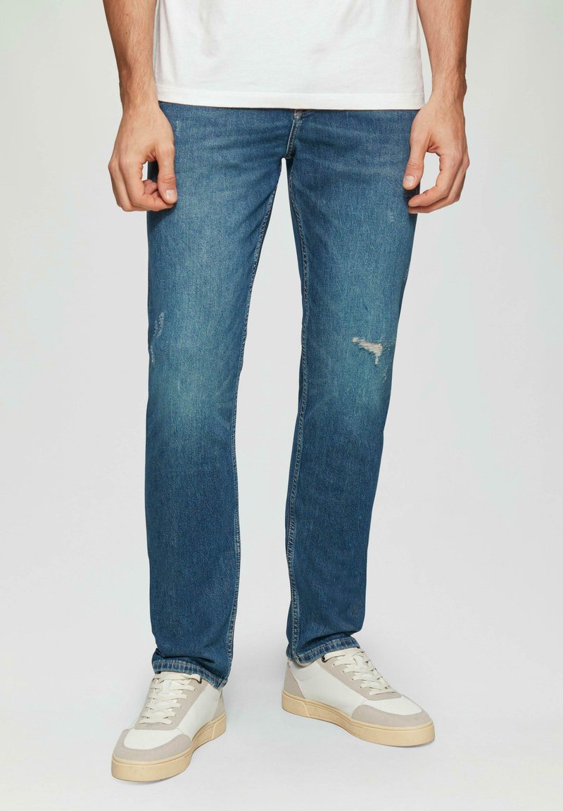 Verbleekte blauwe spijkerbroeken met een rechte pasvorm, voorzien van versleten details op de bovenbenen en gecombineerd met sneakers in neutrale tinten.