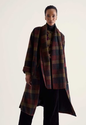 Mujer con un abrigo largo de lana a cuadros y bufanda a juego en tonos rojo oscuro, verde y negro sobre pantalones negros y jersey de cuello alto.