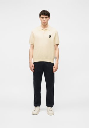 Jeune homme portant un polo crème à manches courtes avec un logo noir, un pantalon noir et des baskets crème, debout devant un fond blanc uni.