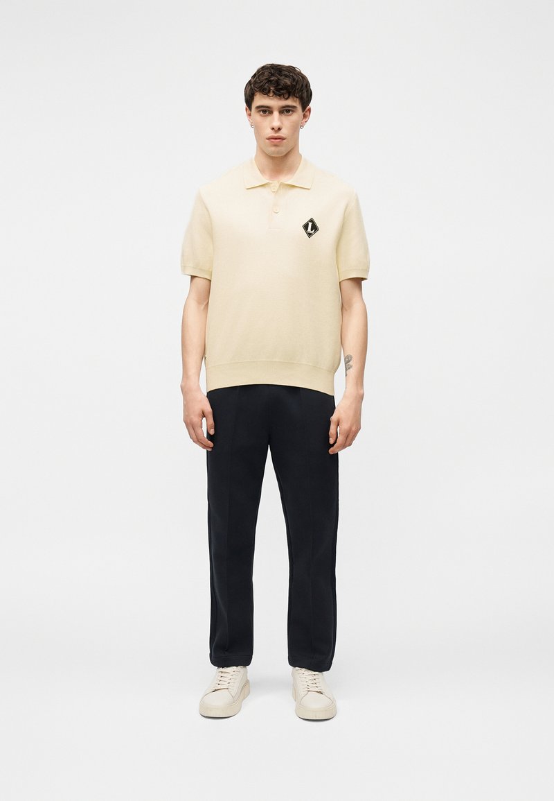 Jeune homme portant un polo crème à manches courtes avec un logo noir, un pantalon noir et des baskets crème, debout devant un fond blanc uni.