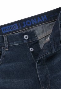 Jeans in denim con un lavaggio scuro, caratterizzati da un comune bottone di chiusura e cuciture a contrasto. L'etichetta del logo riporta "HUGO | JONAH."