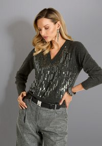 Frau trägt einen dunkelgrauen, mit Pailletten besetzten V-Ausschnitt-Pullover, graue Nadelstreifen-Hose mit schwarzem Gürtel, lange Ohrringe und Armbänder, posiert vor einem grauen Hintergrund.