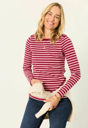 Smiling woman with blonde hair wears a red striped long-sleeve shirt and holds a cream knit sweater around her waist.
