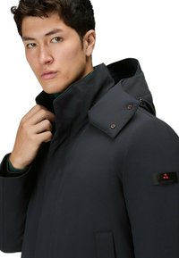 Donkergrijze waterdichte jas met een hoge kraag, capuchon en een schouderdetail met drukknoopsluiting. Heeft een logo-label op de linker mouw.