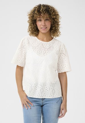 Hvid broderet bluse med korte, flagrende ærmer, der har udskæringsmønstre og en bølget kant. Sammen med lyseblå jeans.