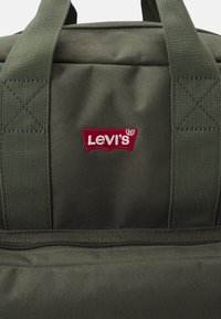 Zielona torba materiałowa z czerwoną naszywką logo Levis. Wytrzymałe paski z przeszyciami krzyżowymi oraz kieszeń zapinaną na zamek u góry. Teksturowana powierzchnia.