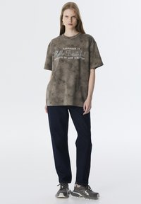 Overmaat olive t-shirt met een vervaagde textuur en witte bedrukte tekst, gecombineerd met donkerblauwe jeans met rechte pijpen en grijze sneakers.