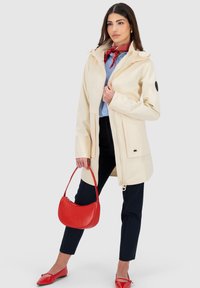 Beige Regenjacke mit Reißverschluss, schwarzem Ellenbogenpatch, kombiniert mit marineblauen Hosen, einer roten Handtasche und roten Flats. Verfügt über eine Kapuze und Seitentaschen.