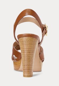 Hnedé kožené sandále na vysokom podpätku s korkovou textúrou, vrstveným podpätkom a s prackou okolo členku, so skríženými popruhmi cez nohu.
