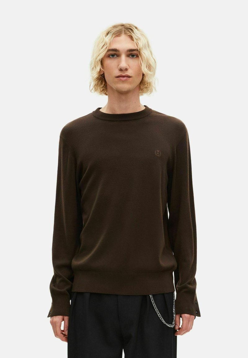 Joven con cabello rizado rubio, vistiendo un suéter de manga larga marrón oscuro y pantalones negros con un accesorio de cadena plateada.