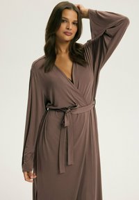 Brązowa sukienka w fasonie kopertowym, wykonana z miękkiego materiału, z długimi rękawami, przewiązywanym w pasie, głębokim dekoltem w serek i bocznymi rozcięciami dla dodatkowego detalu projektowego.