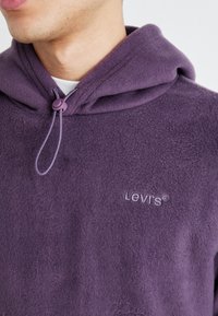 Primo piano di una persona che indossa una felpa viola in pile Levi's con coulisse regolabili e una maglietta bianca visibile al collo.