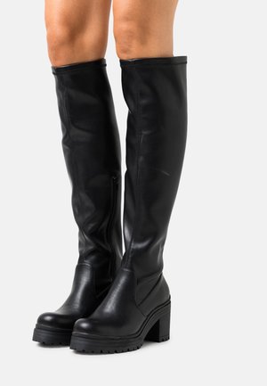 Černé kožené kozačky po kolena s masivními podpatky, nošené na nahých nohách, s postranními zipy a podrážkami s hrubým vzorkem.