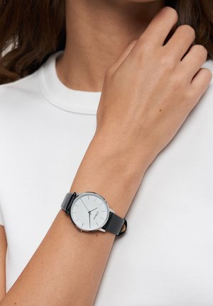 Schwarze Lederarmbanduhr mit silbernem rundem Gehäuse und weißem analogem Zifferblatt. Verfügt über minimalistische Stundenmarkierungen und eine glatte Oberfläche.