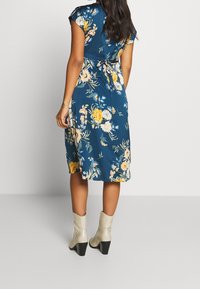 Nainen, jolla on päällään sininen kukallinen midi-mekko, jossa on takana solmittava yksityiskohta, yhdistettynä vaaleanbeigeihin nilkkureihin, seisoo yksinkertaista vaaleaa taustaa vasten.