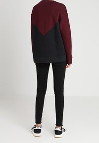 Osoba stojící zády, oblečená v bordovém a černém svetru, černých skinny džínách a černo-bílých teniskách Adidas.