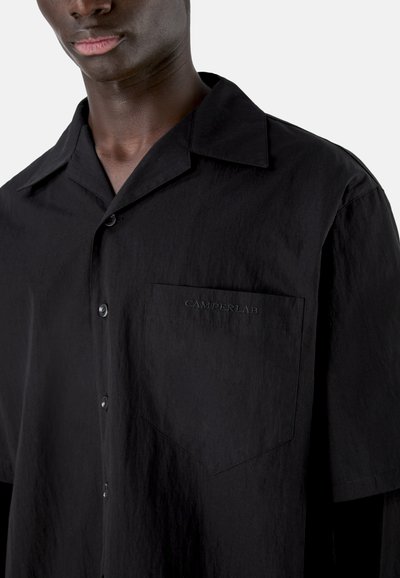 Primer plano de una persona que lleva una camisa negra de manga corta con botones y un bolsillo en el pecho bordado con "CAMPERLAB".