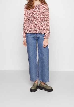 Donna che indossa una blusa rossa floreale a maniche lunghe, jeans blu a vita alta e gamba larga, e mocassini spessi verde oliva, in piedi su uno sfondo neutro.