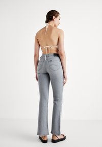Jeans grigi a zampa d'elefante con una texture liscia, dotati di tasche posteriori e una vita nera. La modella indossa un top al collo beige e sandali.