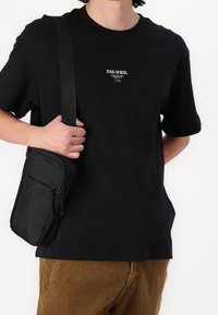 Zwarte katoenen t-shirt met korte mouwen en ronde hals, met witte logodetail. Gecombineerd met een kleine zwarte schoudertas.
