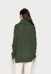 Grüner gestrickter Pullover mit hohem Kragen, lockerer Passform, gerippten Bündchen und Seitenschlitzen, aus strukturiertem Stoff, getragen über hellen Hosen.