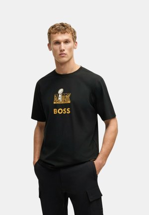 Giovane uomo con maglietta nera con scritte dorate Super Bowl e BOSS, in piedi con le mani in tasca contro uno sfondo semplice.