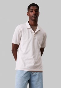 Polo in cotone beige chiaro, con colletto, due bottoni e maniche corte. Abbinato a jeans in denim azzurro.
