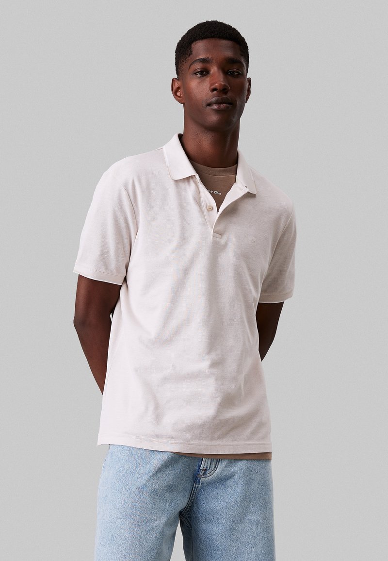 Polo in cotone beige chiaro, con colletto, due bottoni e maniche corte. Abbinato a jeans in denim azzurro.