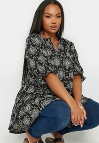 Czarna i biała tunika w kwiatowy wzór o luźnym kroju, z bufiastymi rękawami i dekoltem w serek, połączona z niebieskimi obcisłymi dżinsami i sandałami.