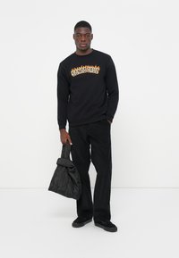 Zwart sweatshirt met "SANTA CRUZ" in vlammenpatroon, gecombineerd met donkere broek en een gewatteerde zwarte tas, staande tegen een witte achtergrond.