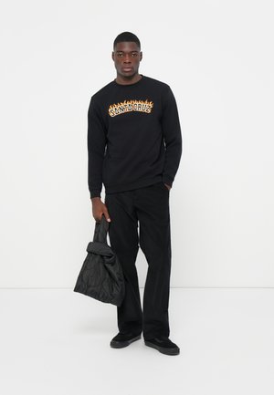 Zwart sweatshirt met "SANTA CRUZ" in vlammenpatroon, gecombineerd met donkere broek en een gewatteerde zwarte tas, staande tegen een witte achtergrond.