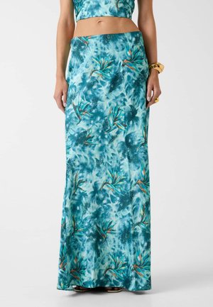 Femme portant une jupe longue maxi à motif floral bleu-vert et un crop top assorti, avec un bracelet et une bague en or à la main droite.