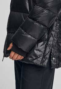 Svart vadderad jacka med glänsande finish, med quiltat mönster, ribbade muddar och sidoförslutning med dragkedja.