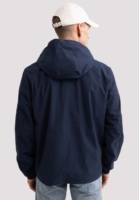 Tumesinine jope kapuutsiga, elastsete varrukatega ja küljetaskuga. Sile tekstuur, vabaaja disain, vaatab tagasi. Kombineeritud heledate teksadega.