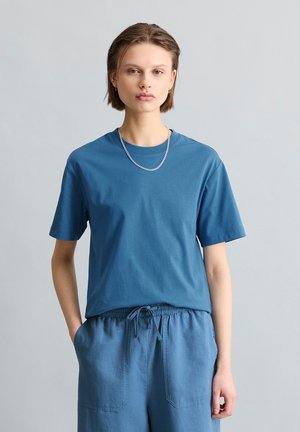 Młoda osoba z krótkimi włosami, ubrana w niebieski t-shirt i pasujące spodnie z sznurkiem, stoi z jedną ręką w kieszeni na jednolitym tle.