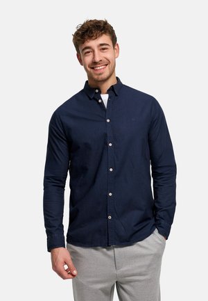 Ung mand smilende, iført en navy blå skjorte med knapper over en hvid undertrøje, med lysegrå bukser, stående mod en ensfarvet baggrund.