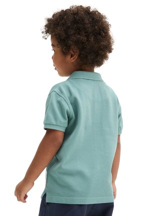 Junges Kind mit lockigem Haar, trägt ein türkisfarbenes Polohemd und dunkle Shorts, blickt nach links vor weißem Hintergrund.