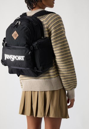 Czarny plecak JanSport z wieloma przegrodami, z diamentowym zdobieniem i logo. Nosi się go na pasku paskowanego swetra i plisowanej khaki spódnicy.