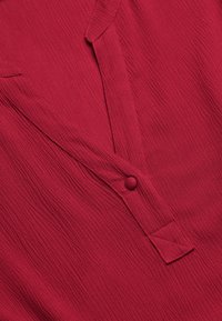 Rode getextureerde blouse met een knoopsluiting, voorzien van een kraag en een diamanten patroon accent bij de sluiting. Details van gekreukt stof zijn zichtbaar.