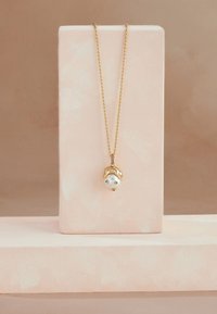 Zelta kaklarota ar pērli kulonā, kas līdzinās sejai, piekārta smalkā ķēdītē. Izstādīta uz gaiši rozā statīva ar reljefainu virsmu.