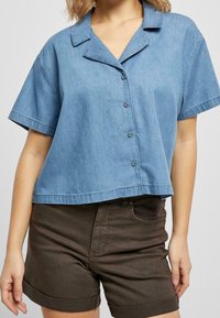 Woman wearing a short-sleeve, light blue button-up cropped shirt with dark brown rolled-hem shorts, standing against a plain background.