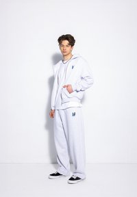 Jasnoszara bluza i pasujące dresy z miękkiego materiału, z małym niebieskim logo na piersi i nogawce. Czarne sneakersy.