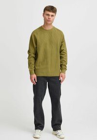 Sweatshirt ribbed de culoare verde măsliniu, cu guler rotund și mâneci lungi, asortat cu pantaloni gri închis. Modelul stă pe un fundal deschis.