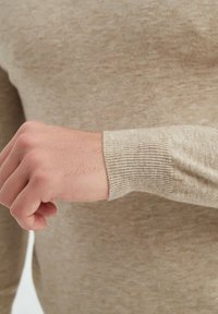 Gros plan sur la main et le poignet d'une personne portant un pull beige à manches longues avec un poignet côtelé.