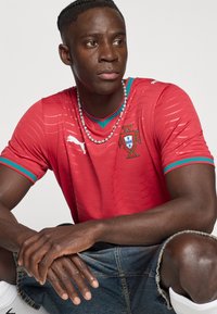 Κόκκινη φανέλα ποδοσφαίρου με μπλε λεπτομέρειες, με σήμα και λογότυπο. Μανίκια κοντά, υφασμάτινη με υφή, φορεμένη με τζιν σορτς και κολιέ με χάντρες.