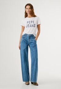 T-shirt bianca con la scritta "PEPE JEANS" in lettere multicolori, abbinata a jeans in denim blu a vita alta e gamba larga, e stivali beige con tacco.