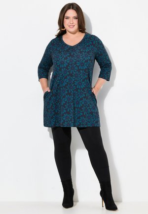 Tunique teal avec motif floral, décolleté en V et manches trois-quarts ; associée à des leggings noirs et des bottines noires. Poches sur les côtés.