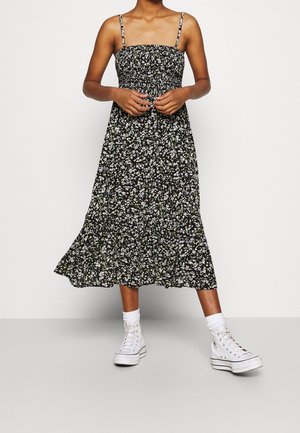 Kvinde iført sort blomster-midi-kjole med tynde stropper, hvide sokker og hvide høje sneakers, står mod en ensfarvet baggrund.