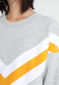 Grijze sweatshirt met een ronde hals en een V-vormig ontwerp met witte en gele strepen. Zachte stoftextuur, casual stijl.