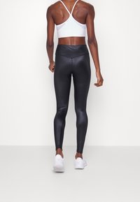 Czarne legginsy o wysokim stanie z błyszczącą fakturą, smukłym krojem i ukośnym szwem z tyłu, zestawione z białymi trampkami.